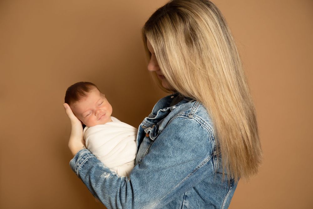 Parent in denim jacket holding swaddled newborn baby against brown background.
