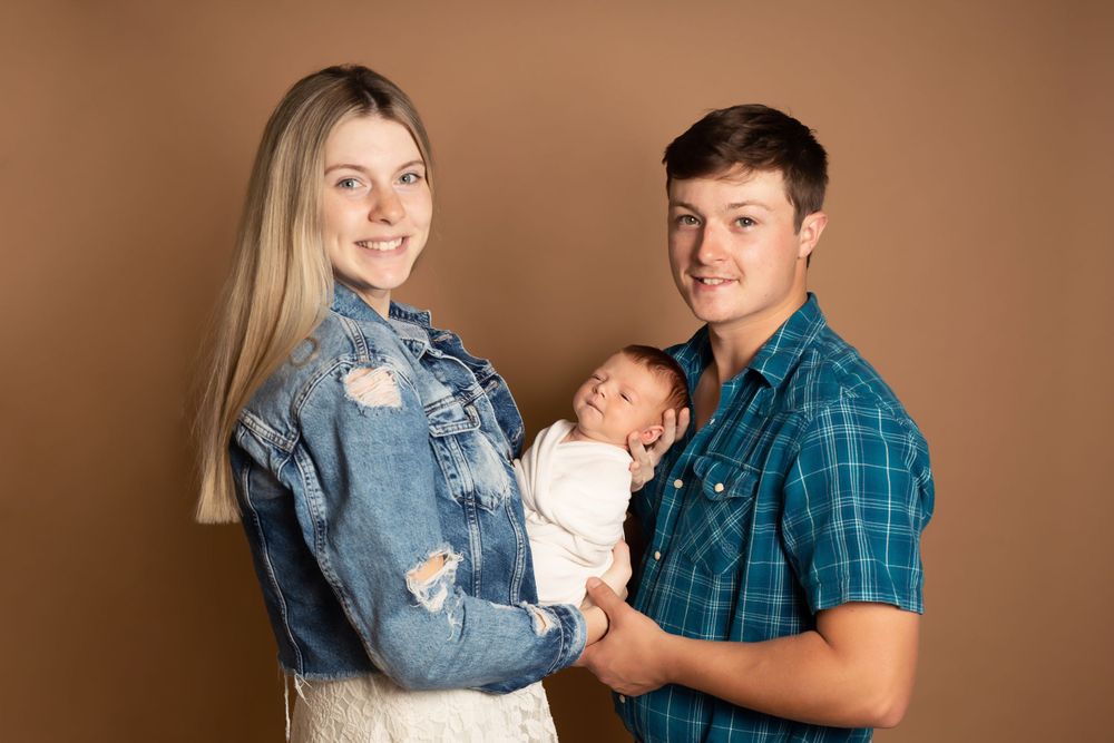Studio portrait with couple in casual attire holding newborn against brown backdrop.