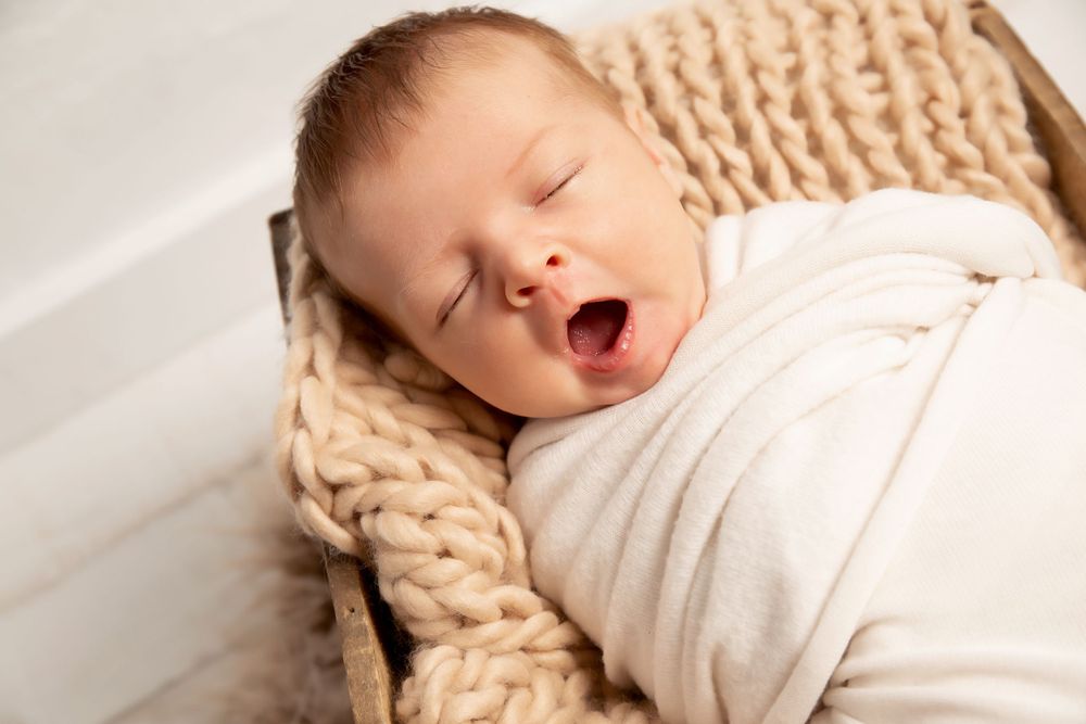 Yawning newborn baby swaddled in white on knitted blanket.