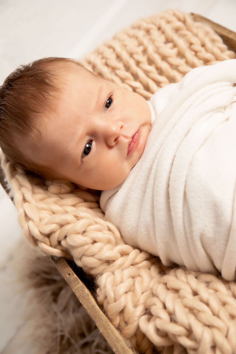 Baby wrapped in white fabric lying on chunky knit blanket with soft lighting.