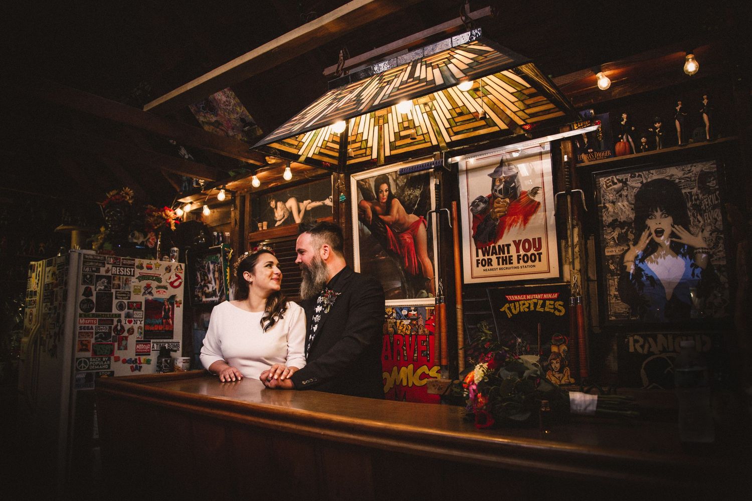 Couple poses at vintage bar with rustic decor and dim atmospheric lighting.