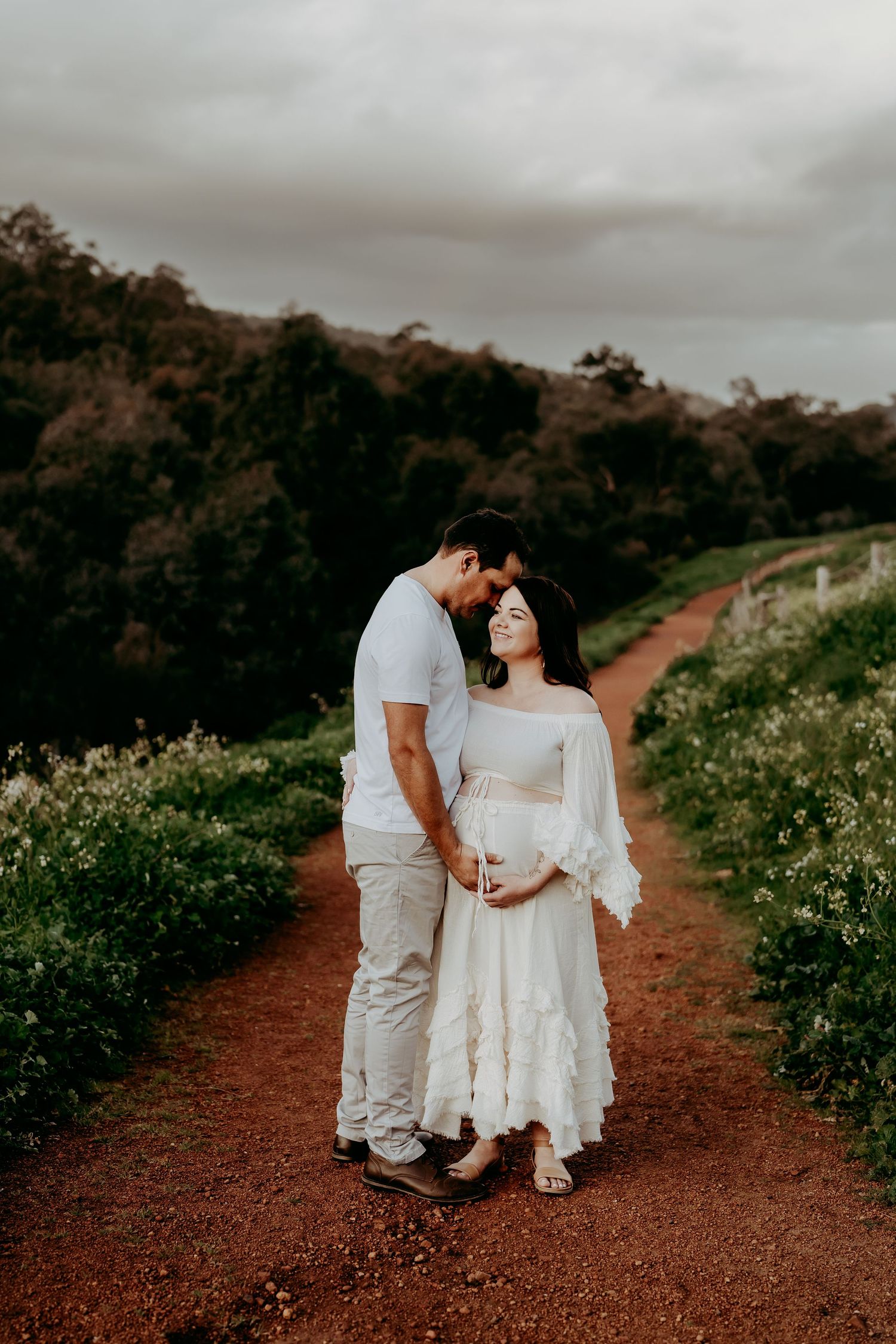 A couple in light colored clothing poses together on a rustic red dirt path surrounded by nature.