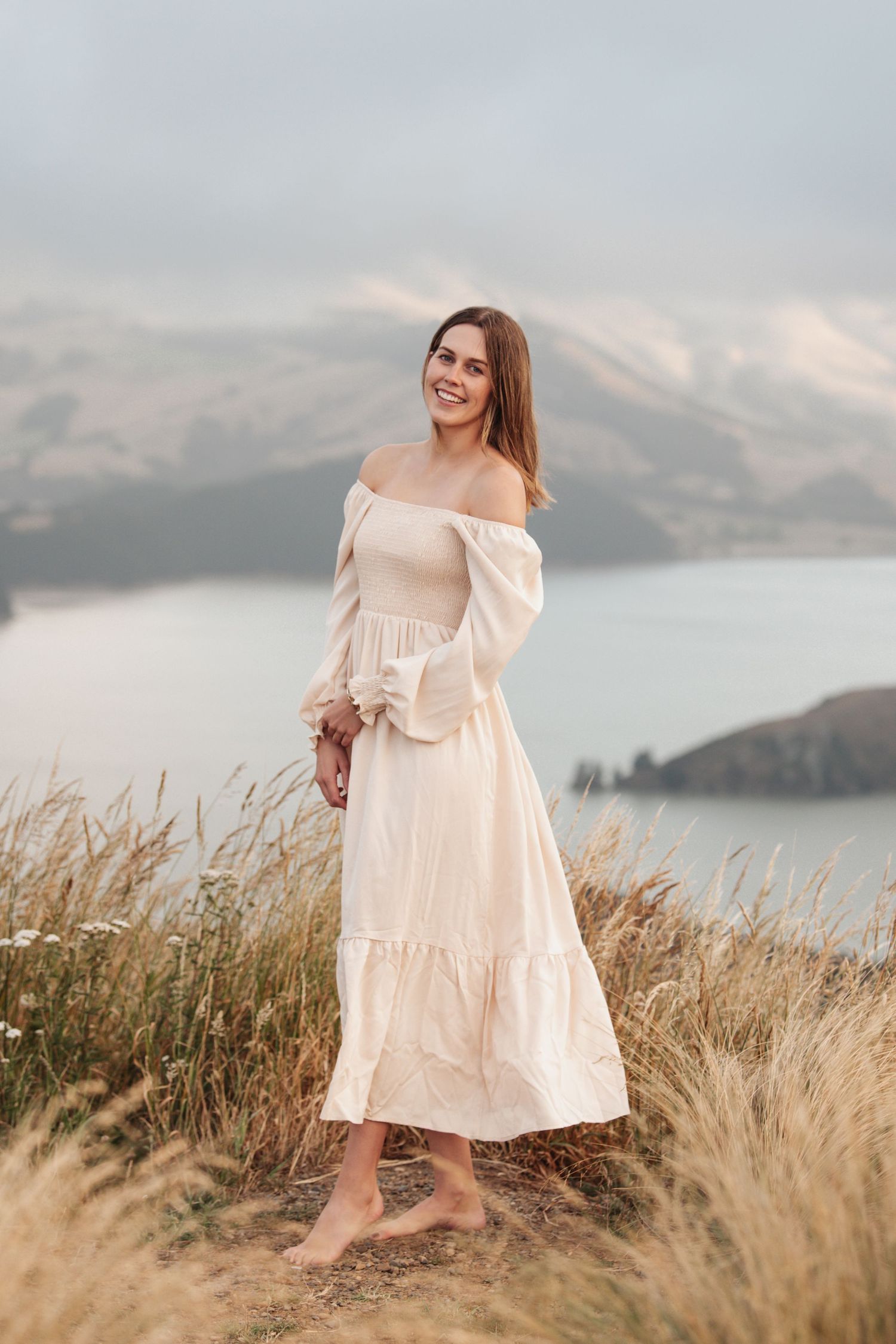 Person in cream off-shoulder dress standing on coastal hillside with mountain lake view.