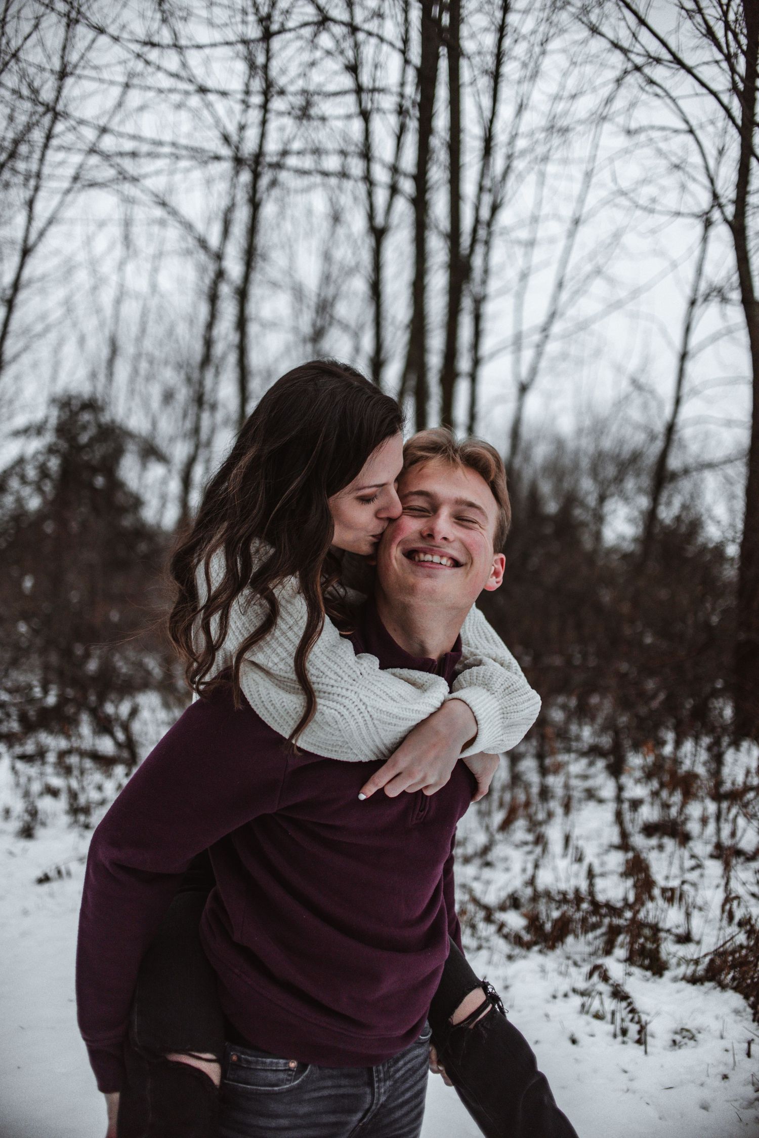 A candid winter engagement photo showing a playful piggyback ride in a snowy forest setting.