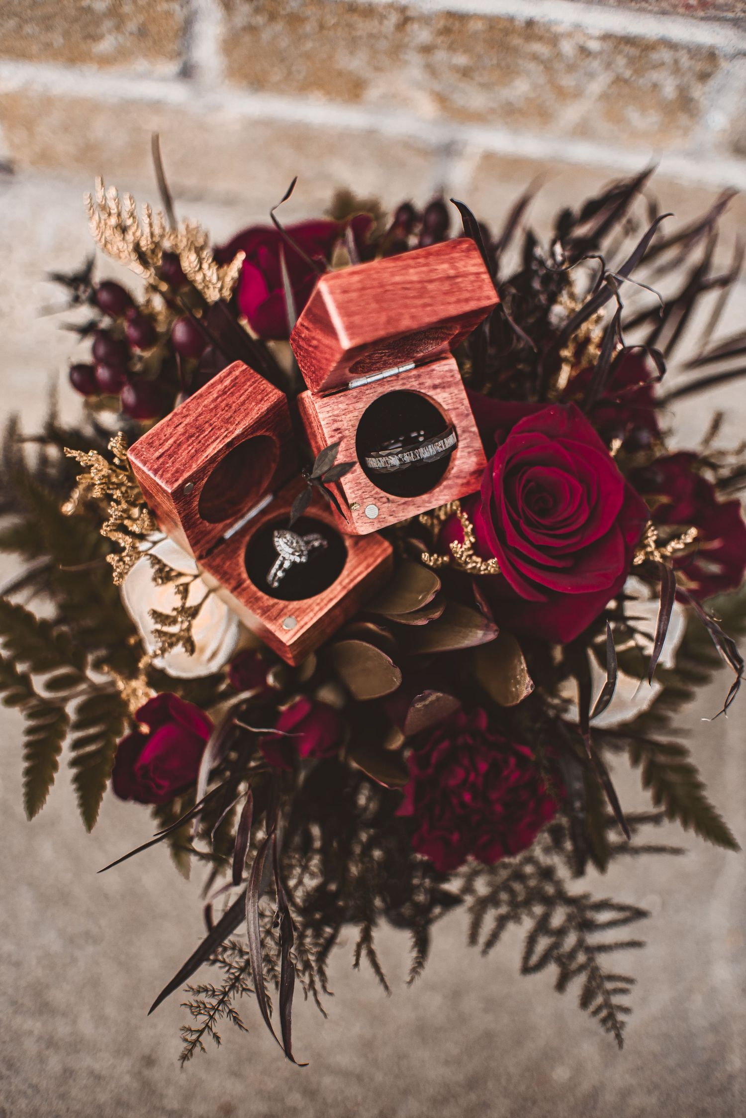 Wedding rings displayed in coral boxes atop a dramatic floral arrangement with deep red roses.