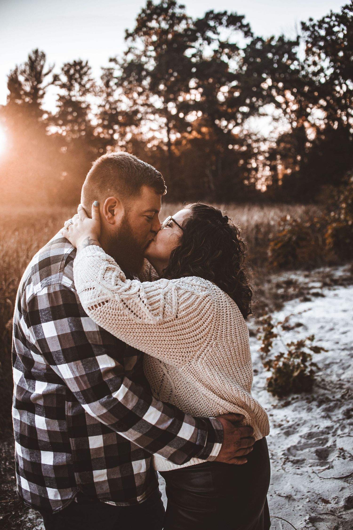 A couple embraces in a snowy setting during sunset wearing cozy winter attire.