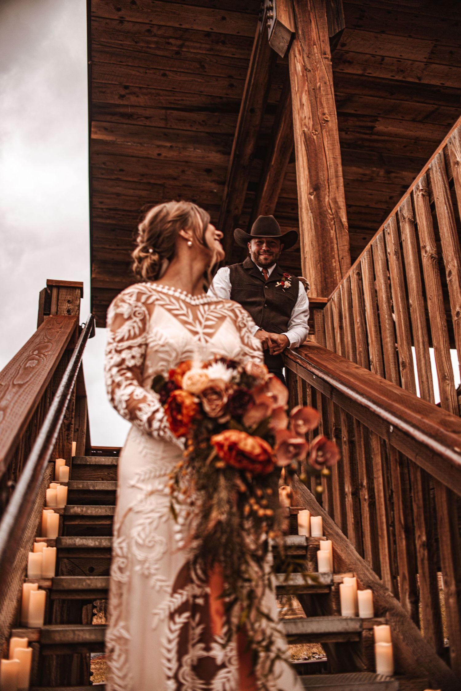 A bride in a detailed lace gown ascends wooden stairs holding an autumn bouquet.