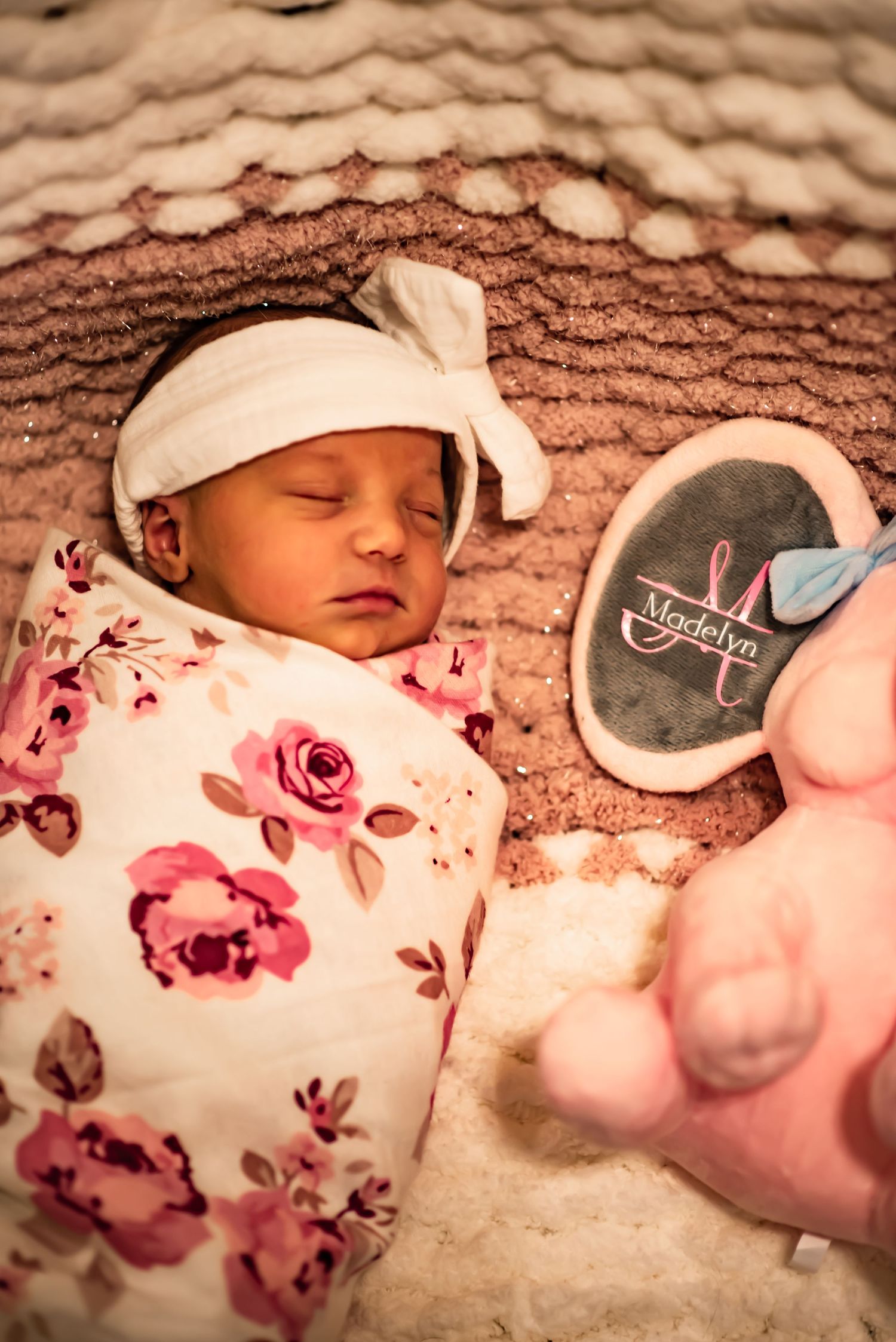 Sleeping newborn wrapped in floral swaddle with white headband next to plush toy.