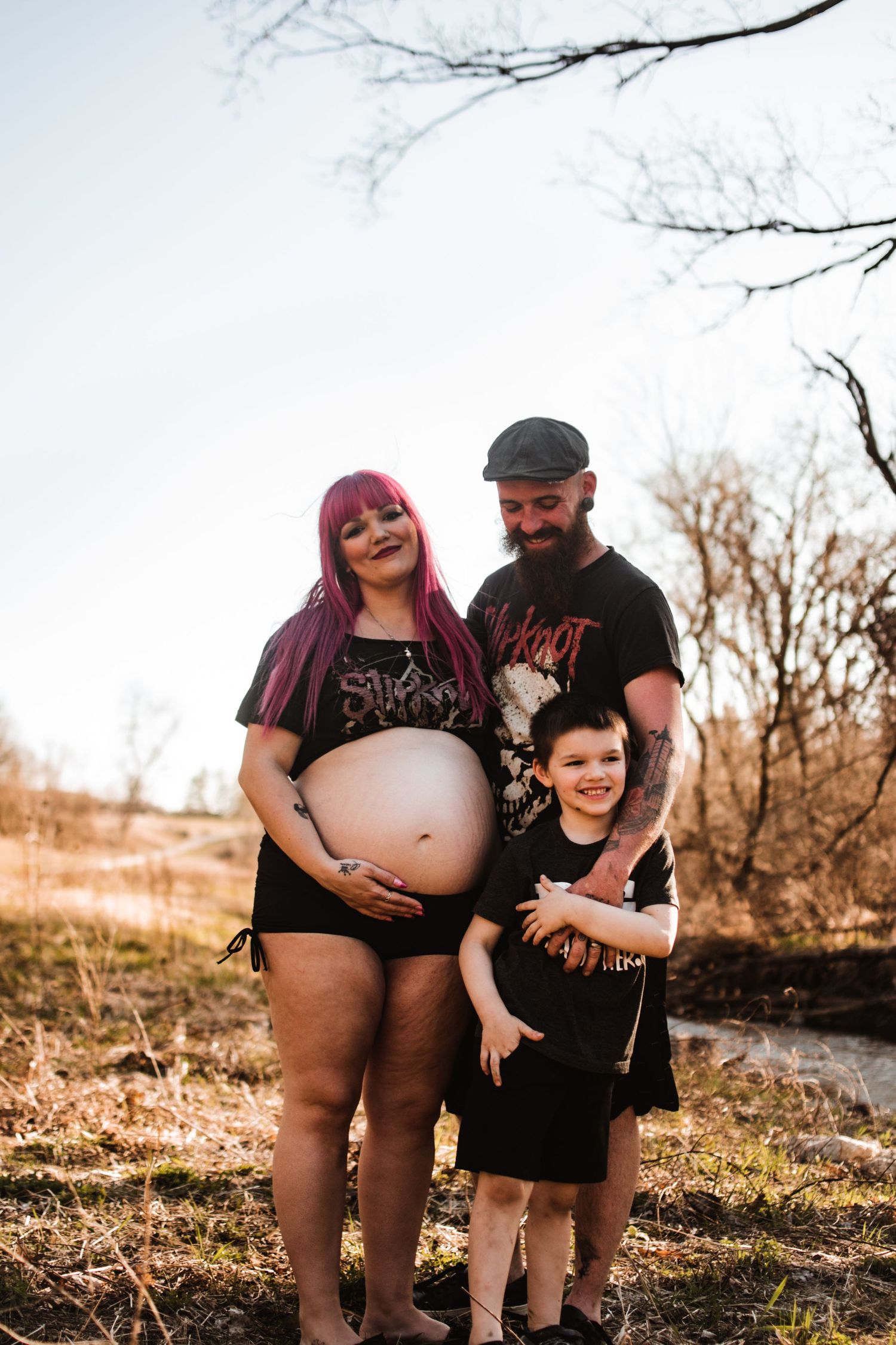 Expecting family poses outdoors in casual clothing against a natural autumn backdrop.
