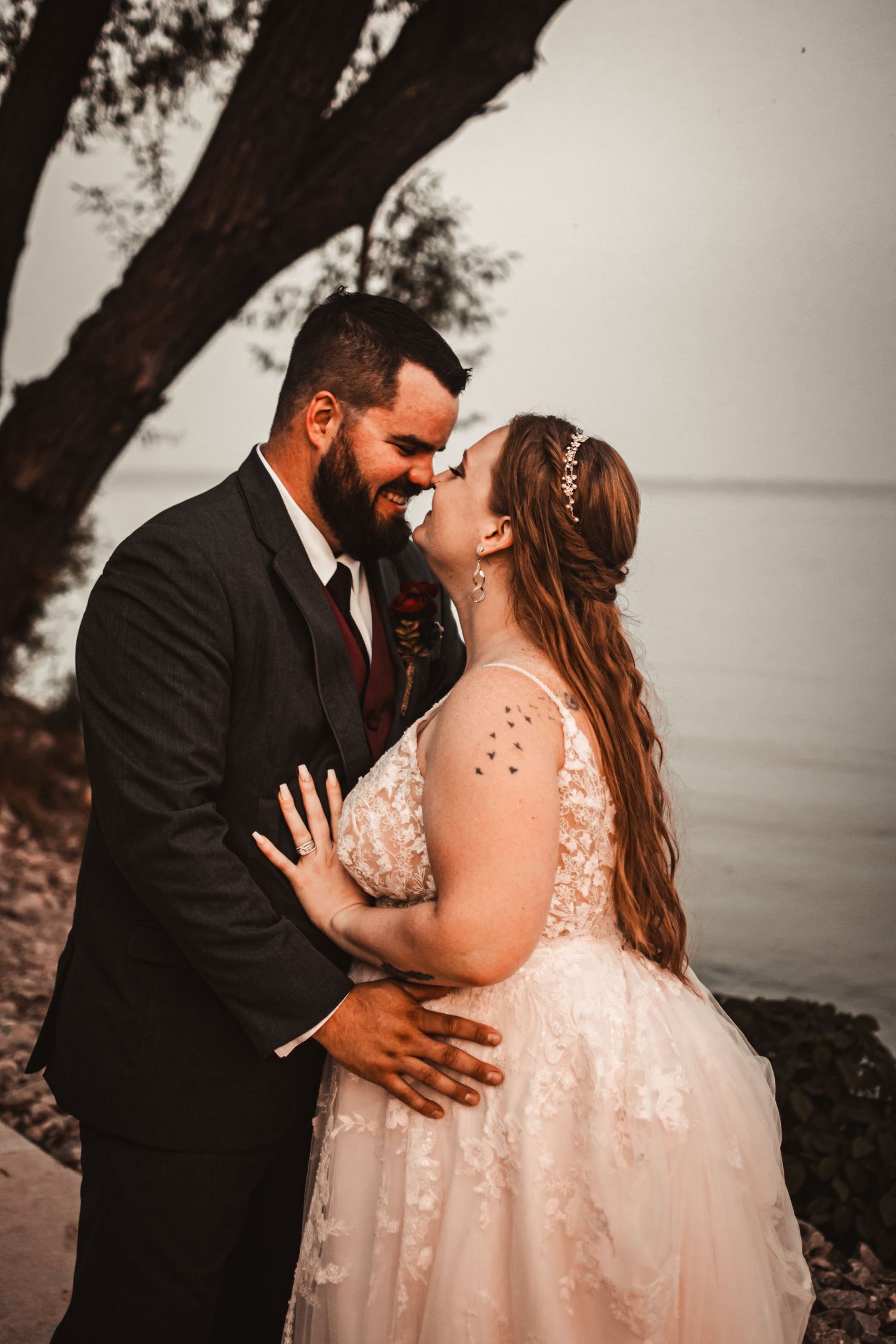 A romantic wedding photograph showing a couple embracing under a tree at sunset by a waterfront.