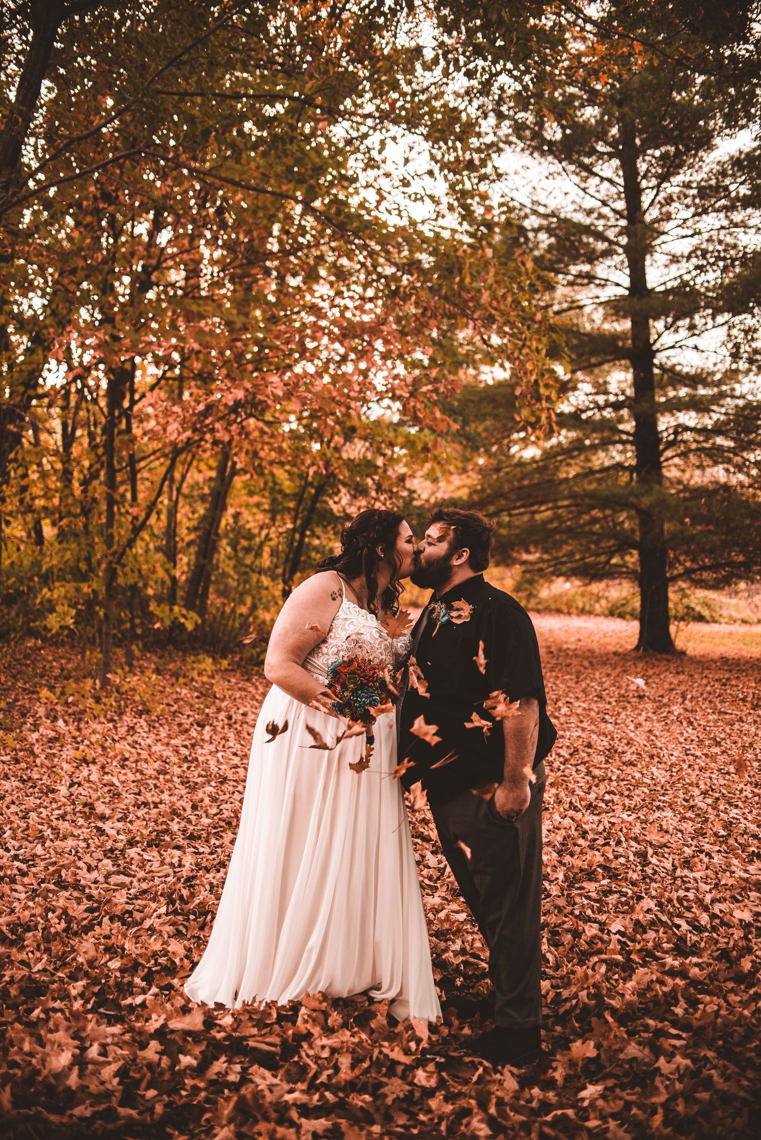 A romantic fall wedding photo shows a couple embracing on a carpet of autumn leaves in a forest at sunset.