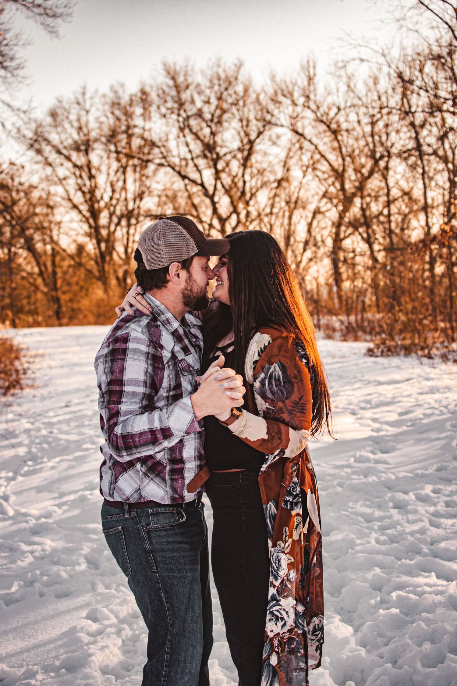 A couple shares an intimate moment in the snow during golden hour with bare trees in the background.