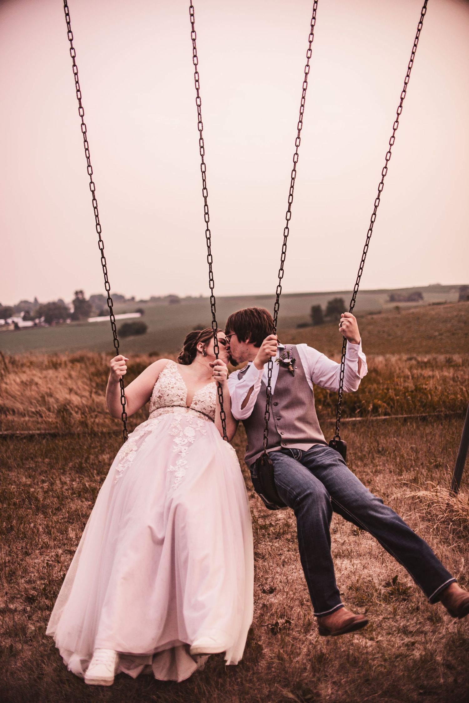 A couple shares an intimate moment while sitting together on swings in a rustic field.