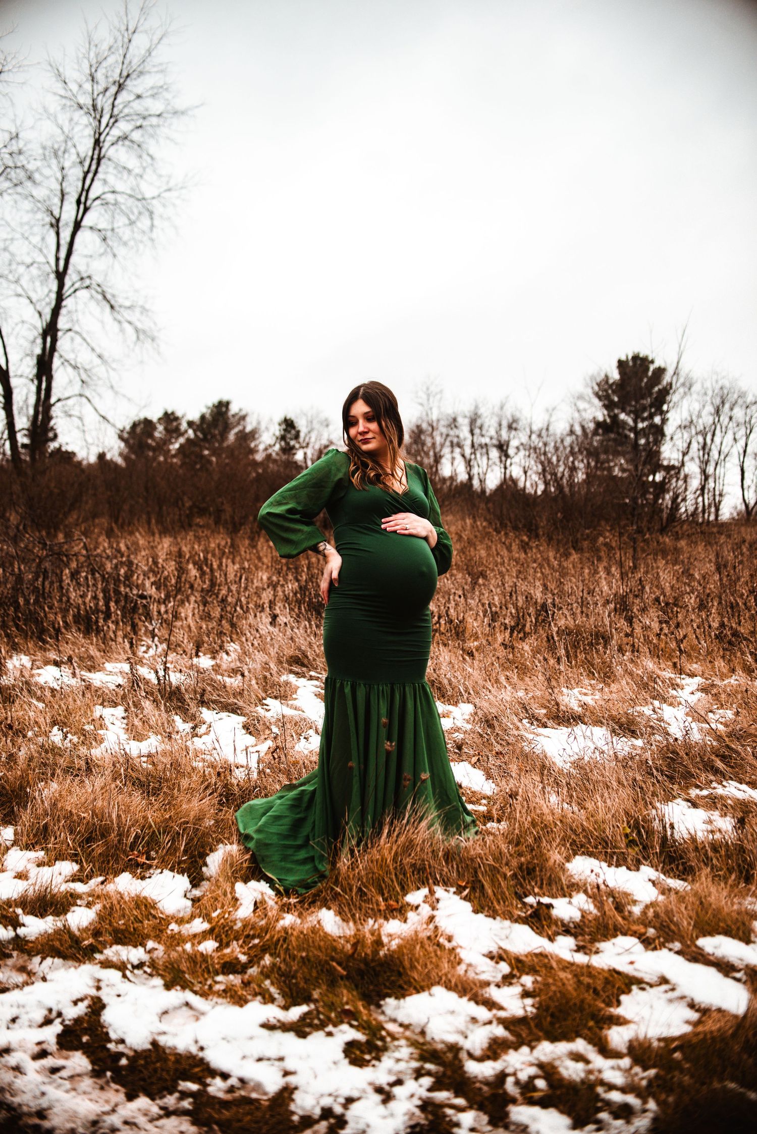 A maternity photo of a woman in an elegant green dress standing in a snowy winter field.