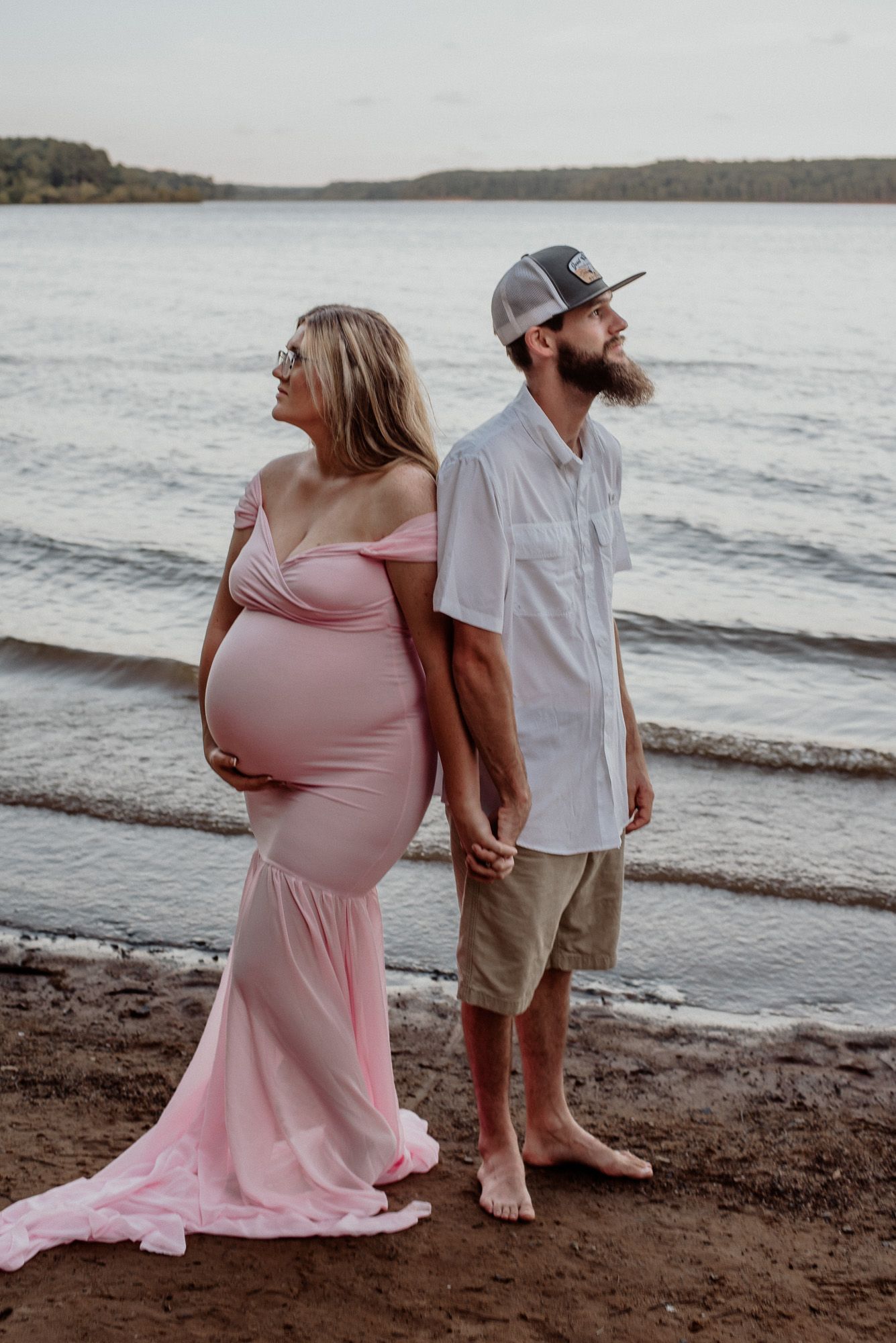 Expecting couple standing on beach at sunset, wearing pink dress and casual attire.