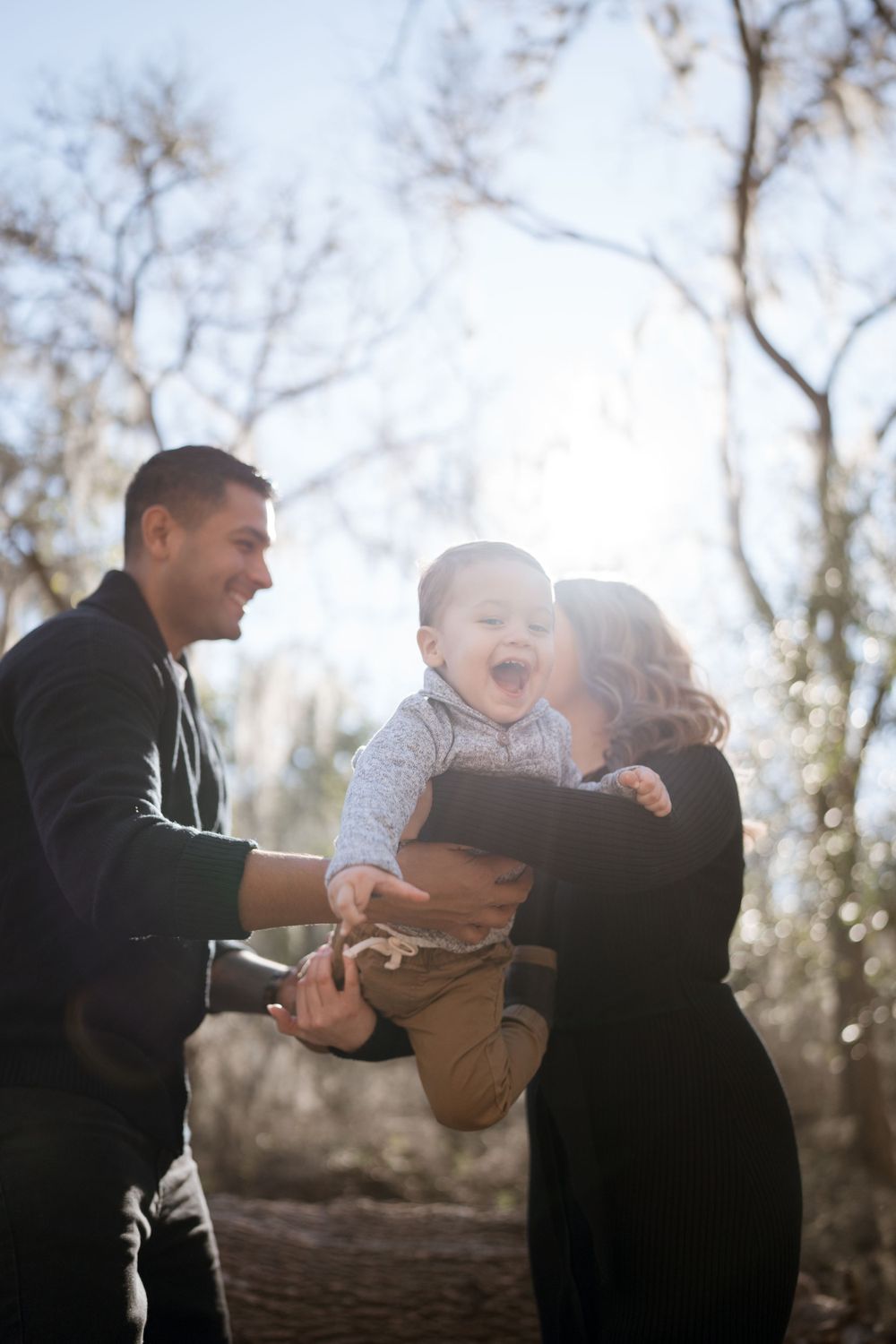Happy family plays outside in nature as parents lift their laughing child together on sunny day with trees and sunlight.