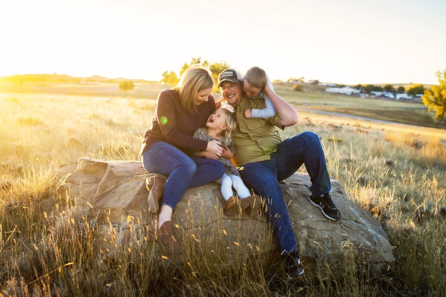 Family relaxing together on rock during golden hour with sunlit field in background.
