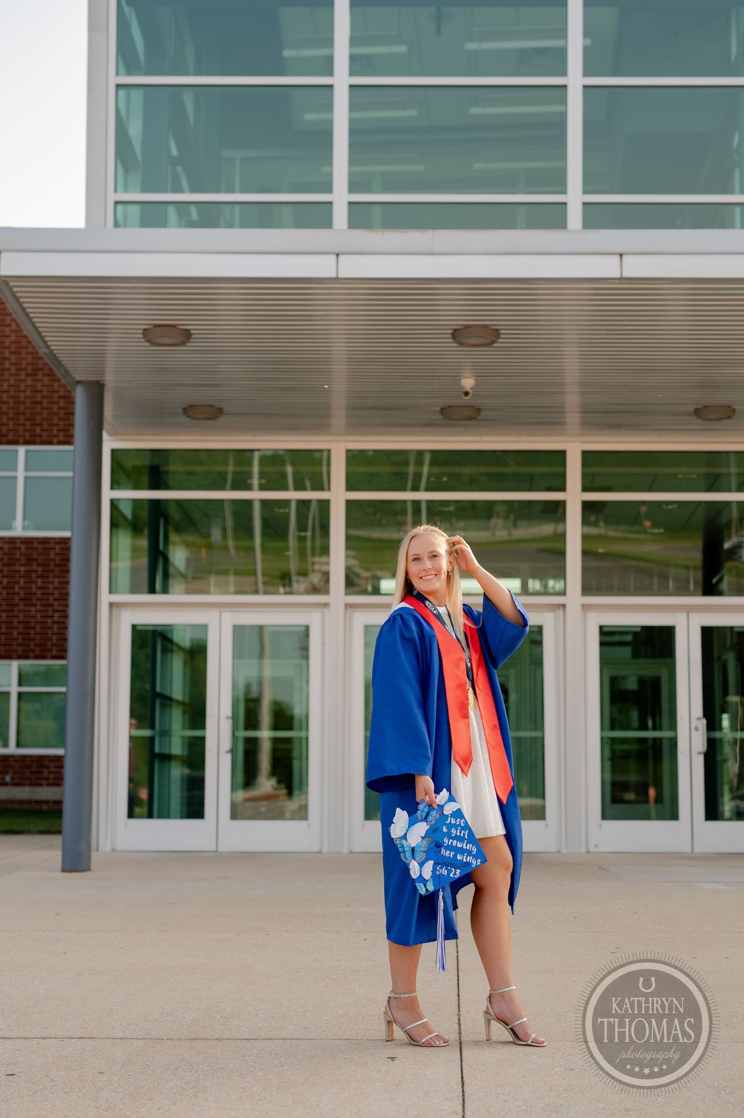 Graduate in blue graduation attire poses confidently outside modern school entrance.
