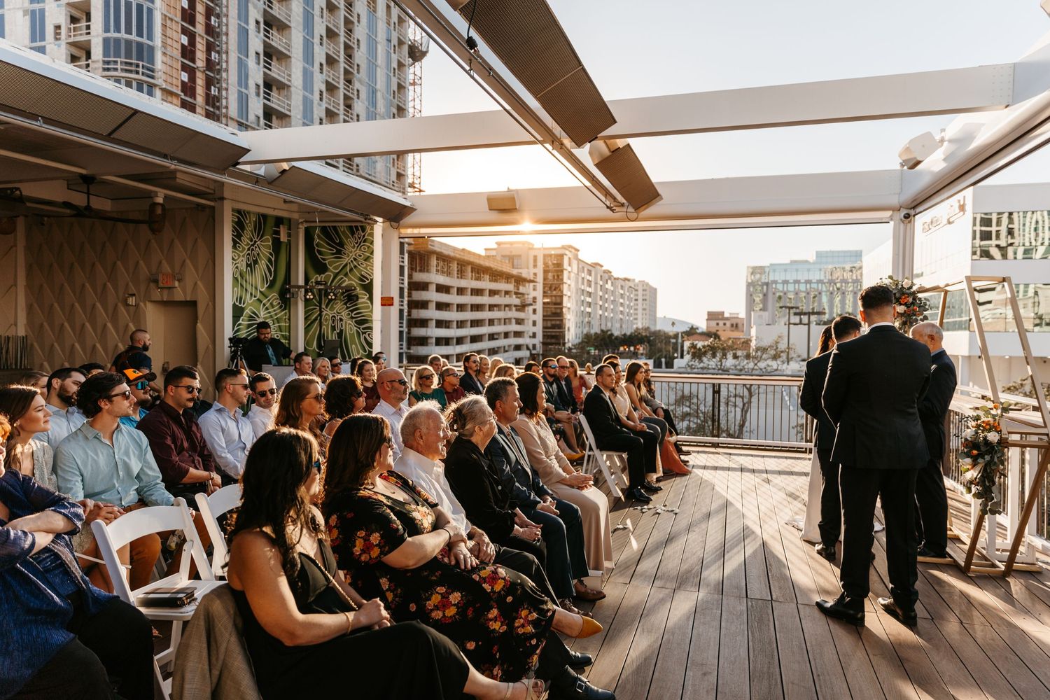 Guests seated at rooftop wedding ceremony at sunset with city skyline backdrop and officiant at altar.