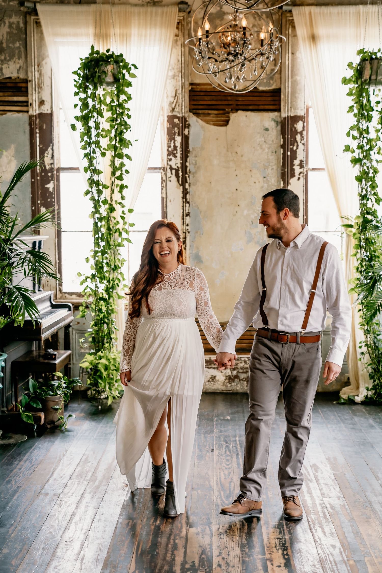 People in romantic attire walking together in vintage room with hanging greenery and curtains.