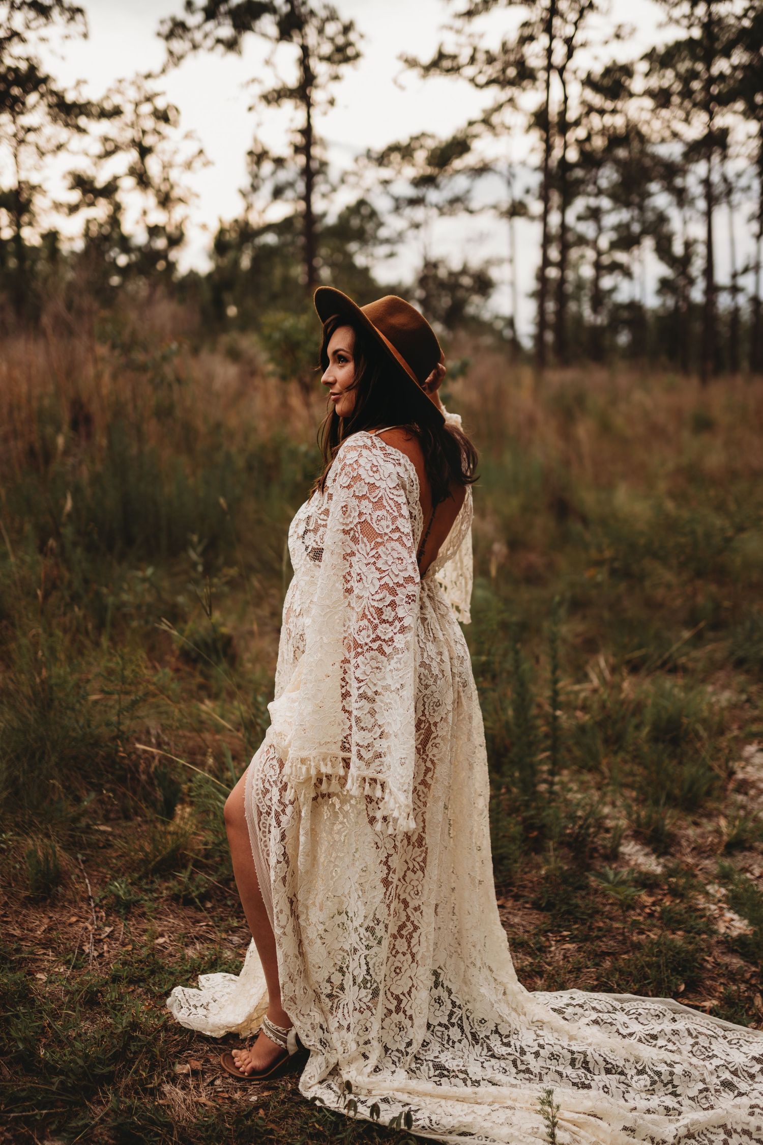 Bohemian maternity portrait in white lace dress and hat against wooded backdrop at sunset.