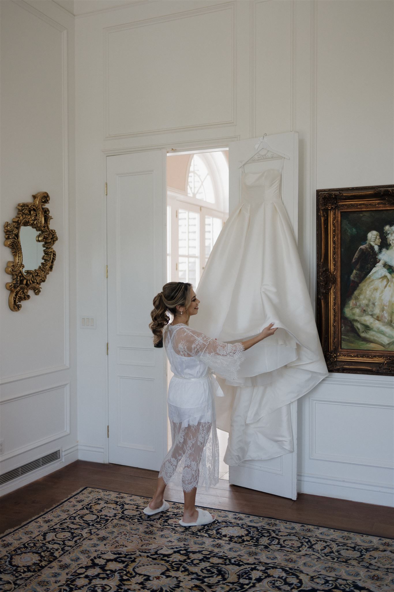Elegant wedding dress hangs in bright white room with ornate decor including baroque mirror and vintage rug.