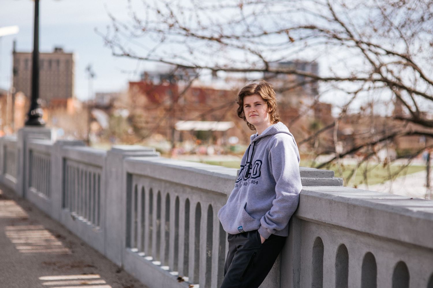 A casual figure in a gray hoodie stands by a concrete railing in an urban setting.