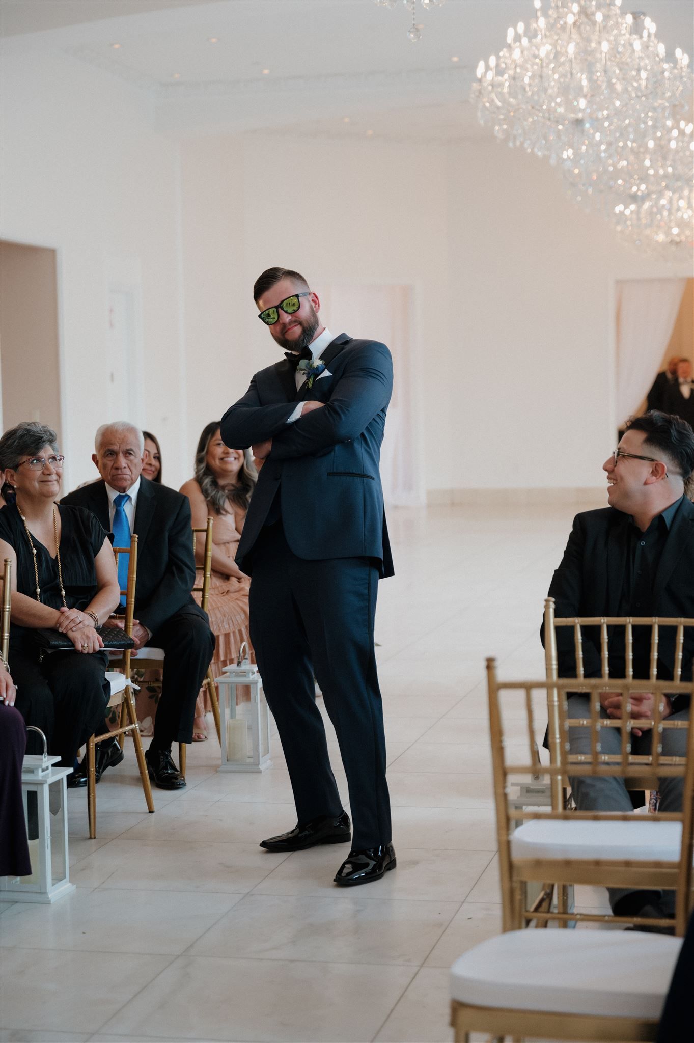 An elegantly dressed guest stands in a bright venue with chandeliers and gold chairs during a formal event.