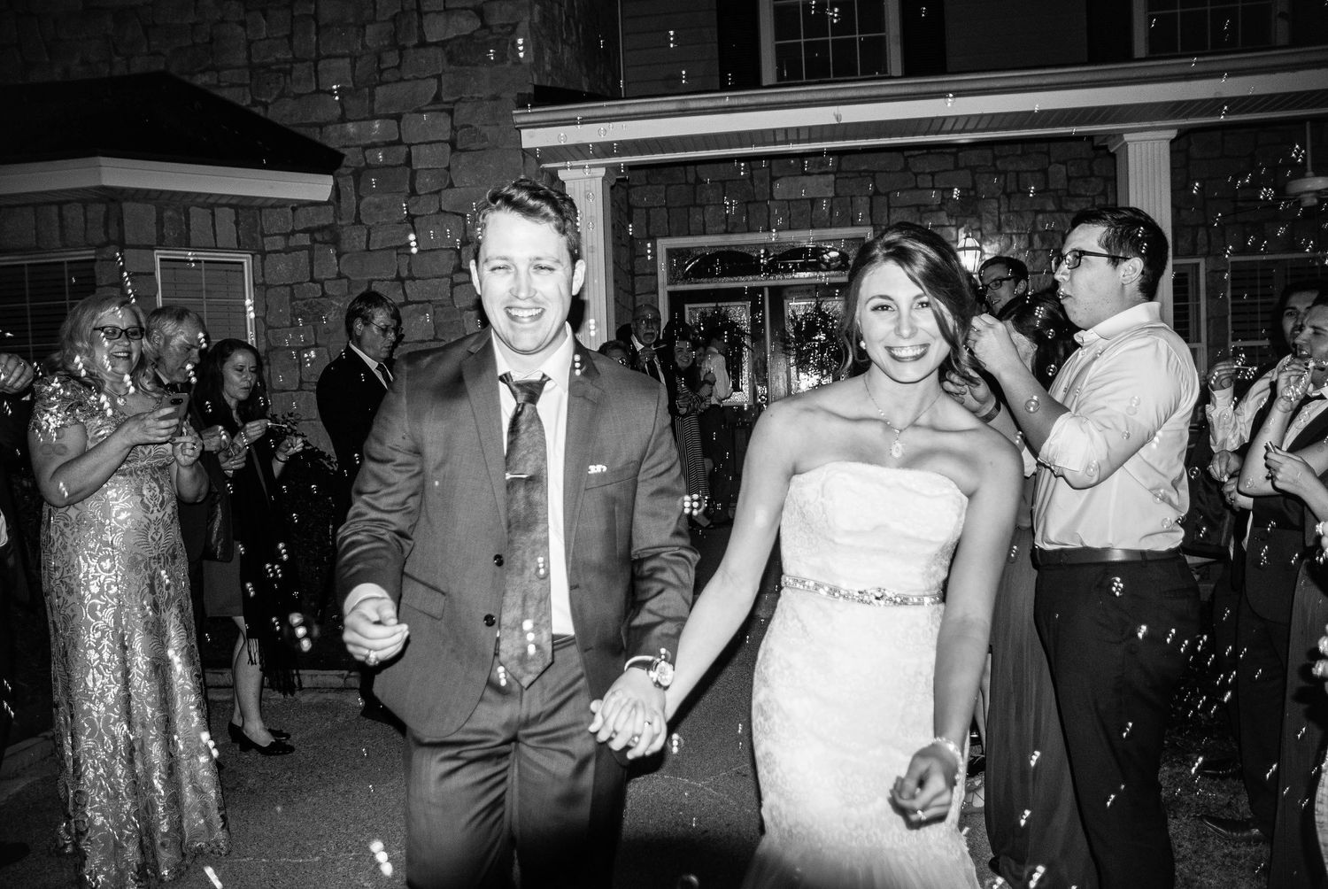 Happy bride and groom walking hand in hand through confetti at their wedding reception in black and white photo.