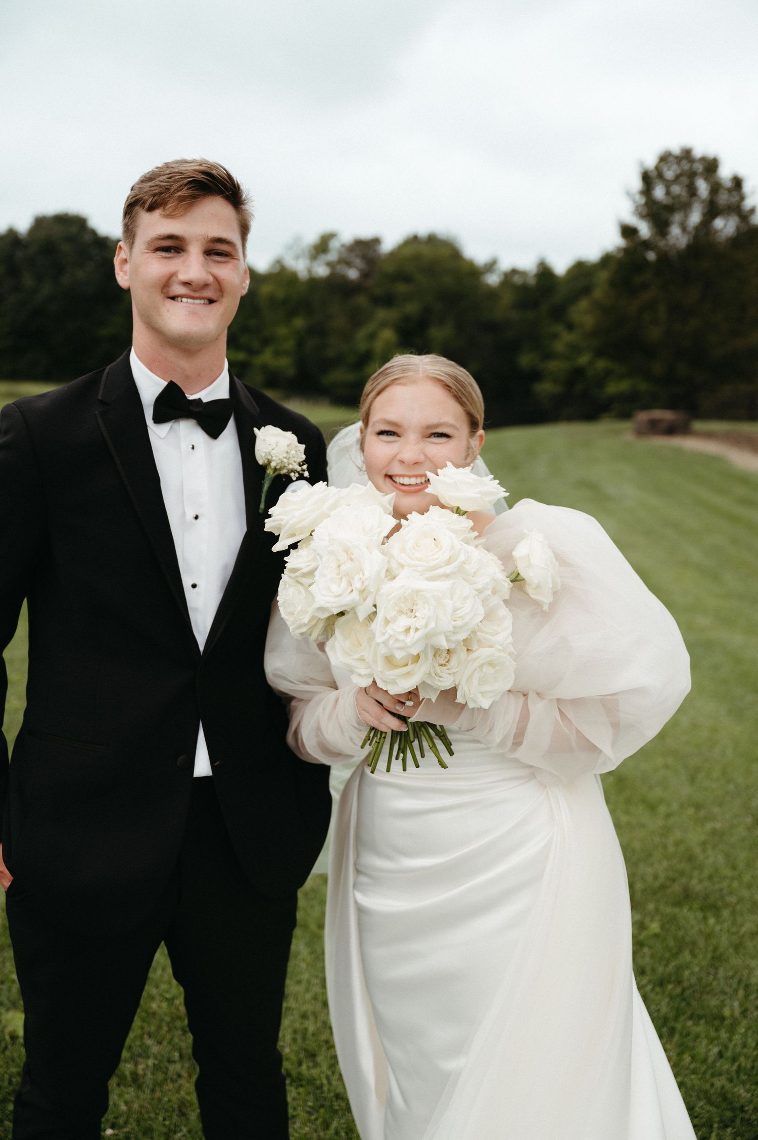 A wedding couple poses outdoors holding a large bouquet of white roses.