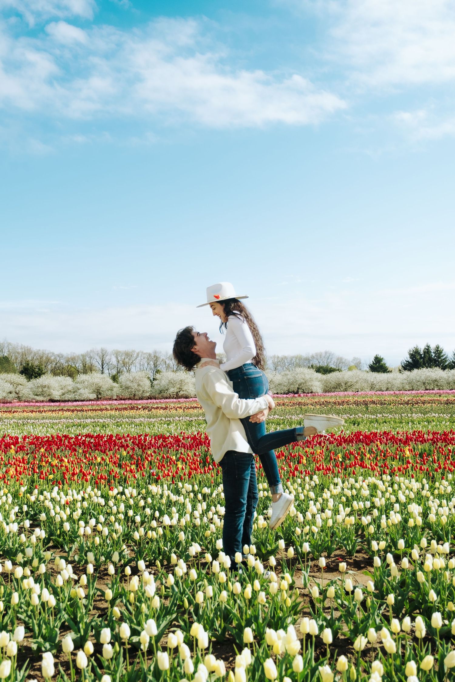 Playful couple dancing and twirling among rows of red and white tulips in a sunny flower field.