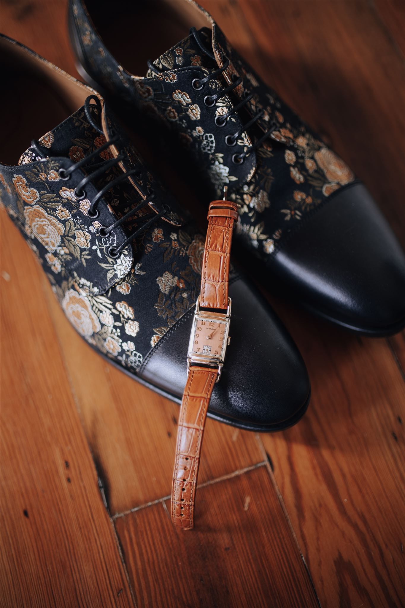Floral patterned dress shoes and brown leather watch on wooden floor.