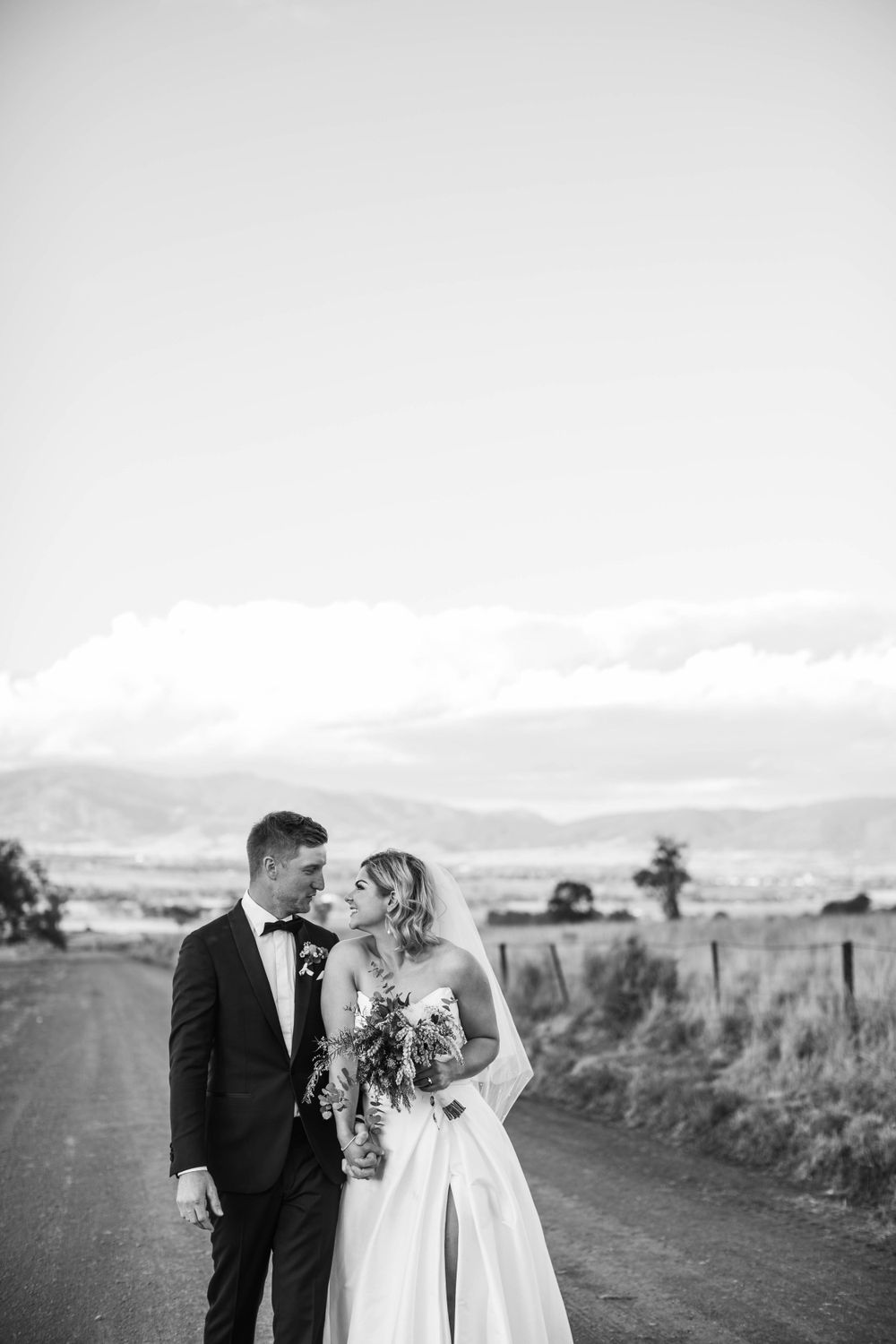 Wedding couple standing on a dirt road with mountains in the background.