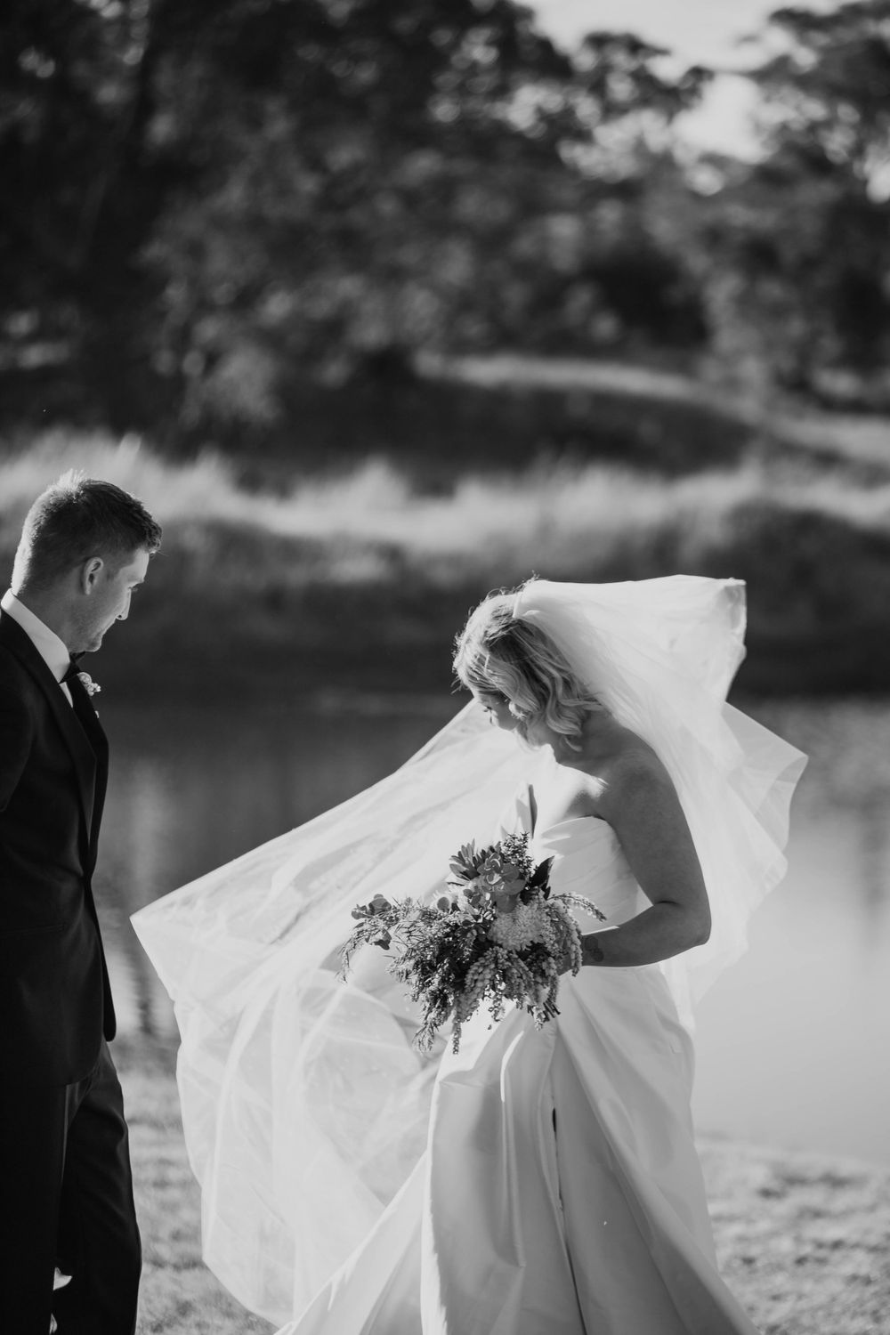 A bride in a flowing white dress and veil stands with her groom by a lake in a black and white photo.