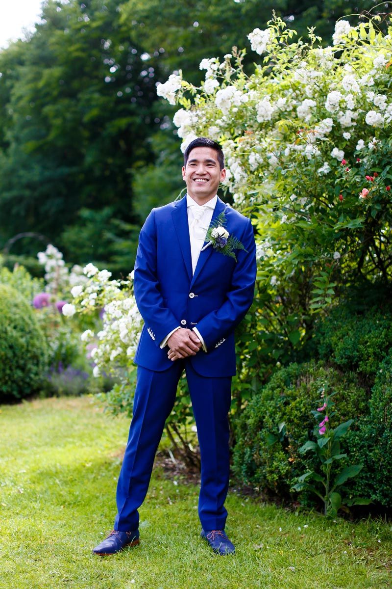 Groom in royal blue suit poses in garden with blooming white roses.
