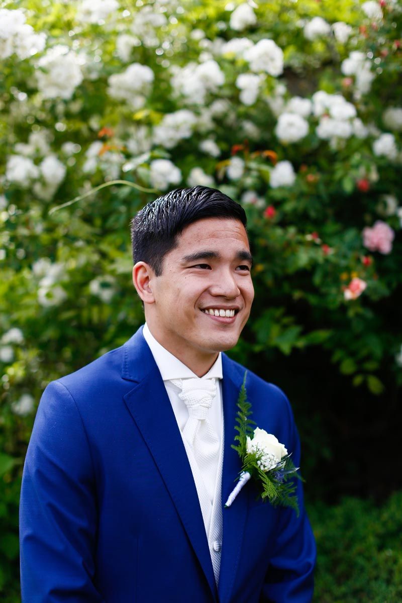 Portrait of groom in navy suit with white boutonniere against rose garden backdrop.