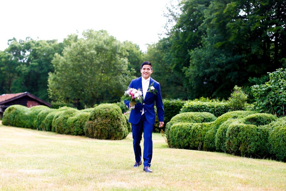 Groom in bright blue suit walking along manicured garden hedges.