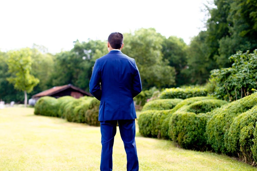 Back view of groom in royal blue suit standing by formal garden hedges.