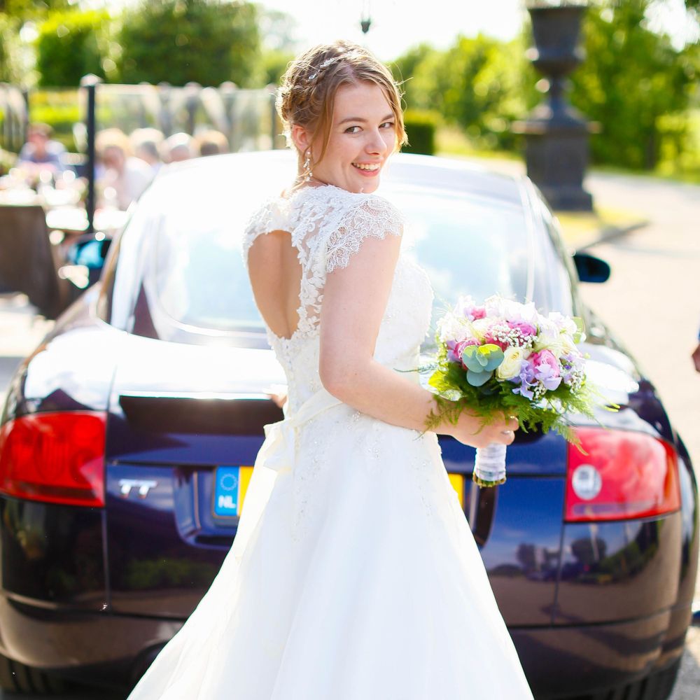 A bride in a lace-detailed dress poses with her bouquet next to a car.