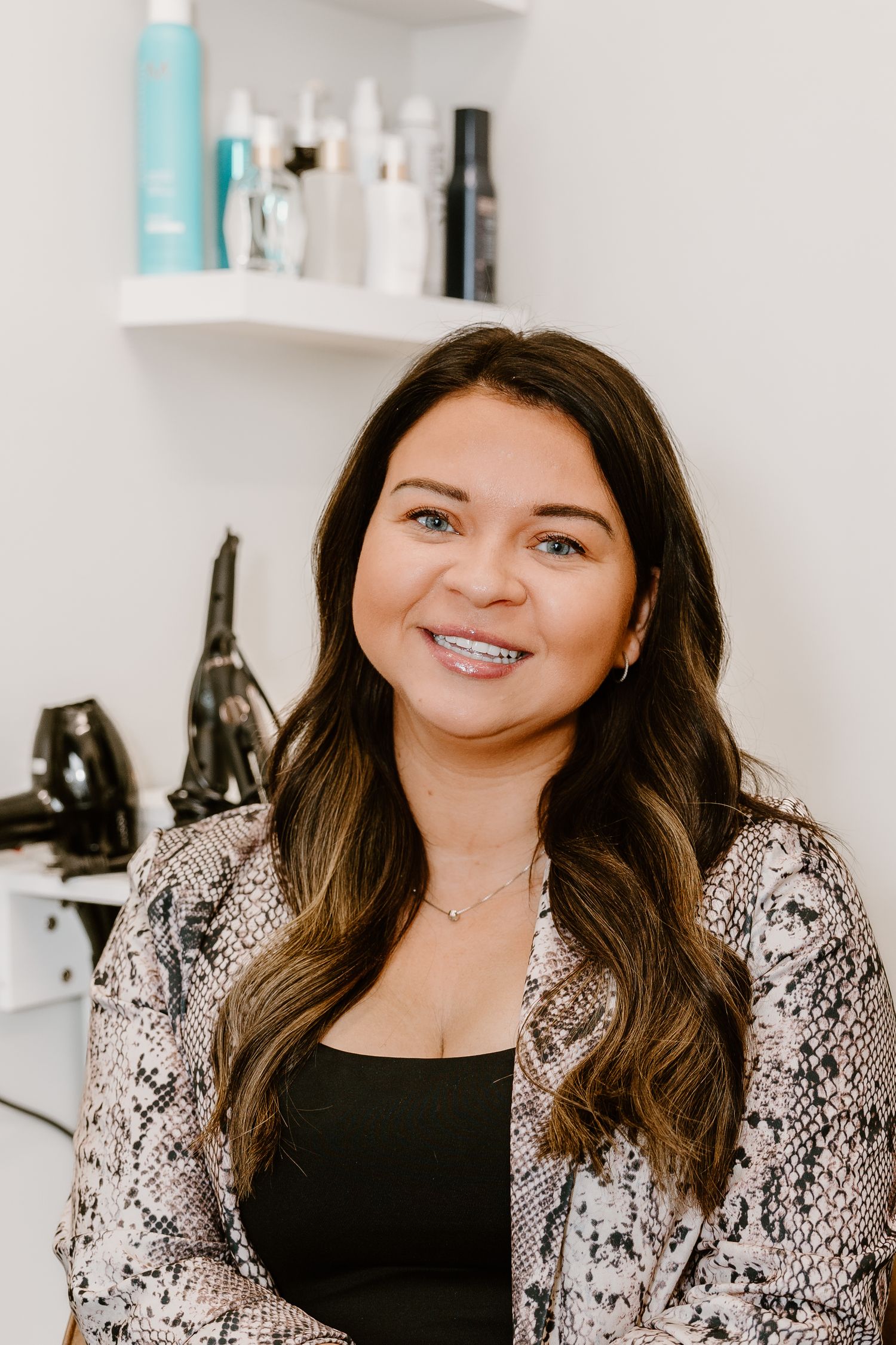 A hairstylist in a snakeskin print blazer poses in a salon with product shelves in background.