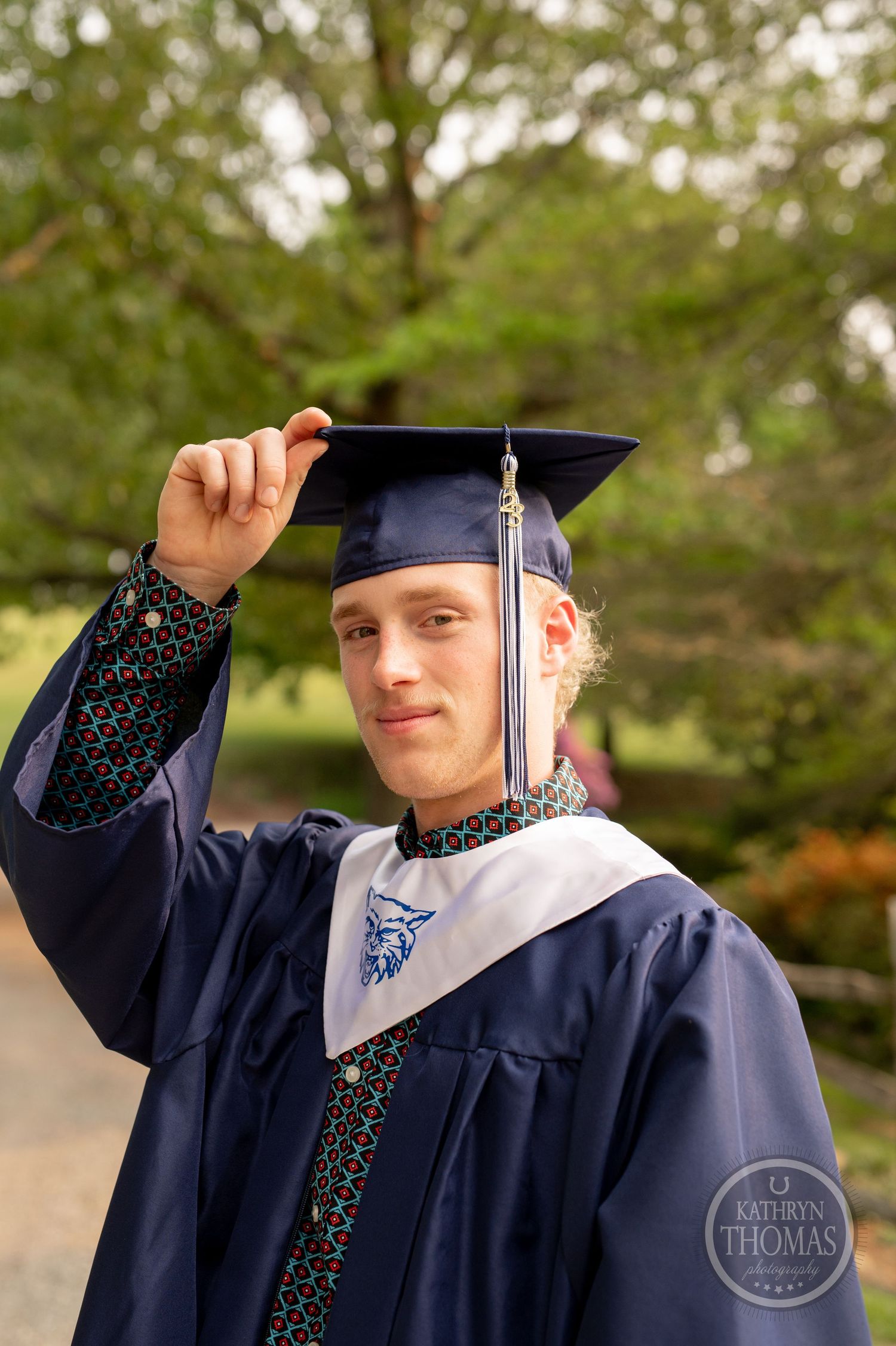 Graduate in navy blue regalia holds cap tassel while posing outdoors.