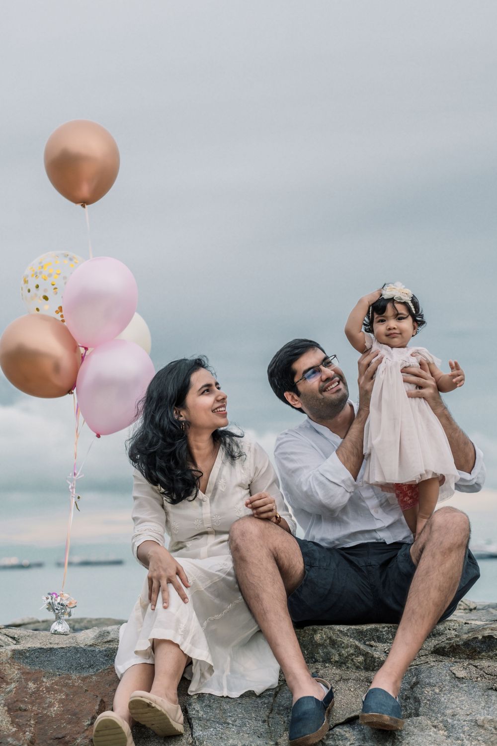 Family celebrates with pink and gold balloons while sitting on coastal rocks.