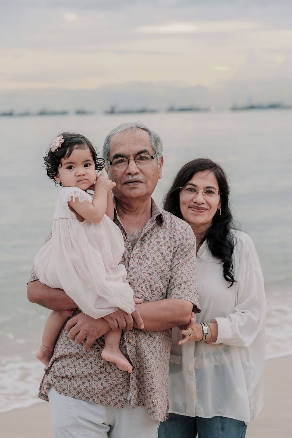 Family of three poses together on beach at sunset in casual white outfits.