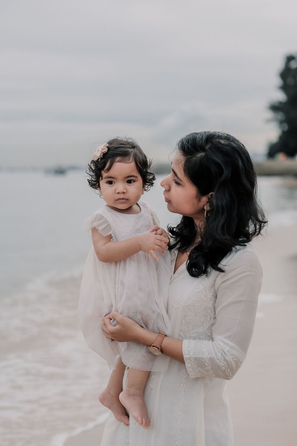 Parent holding a toddler in a white dress on a peaceful beach.