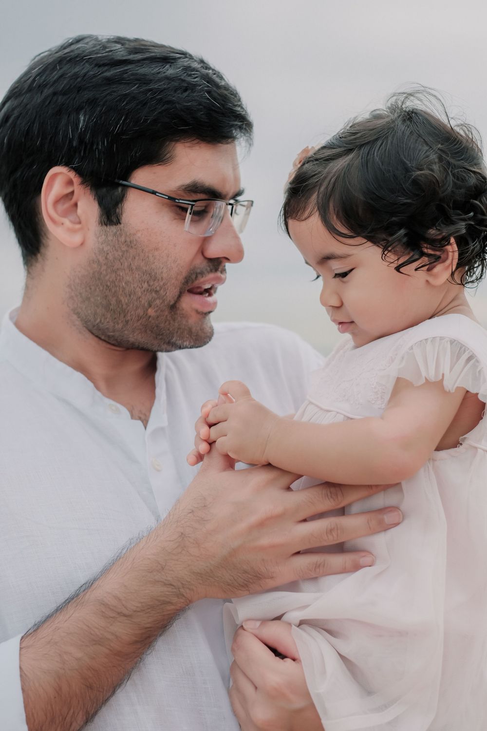 Parent and toddler share a tender moment in matching white outfits against a light background.