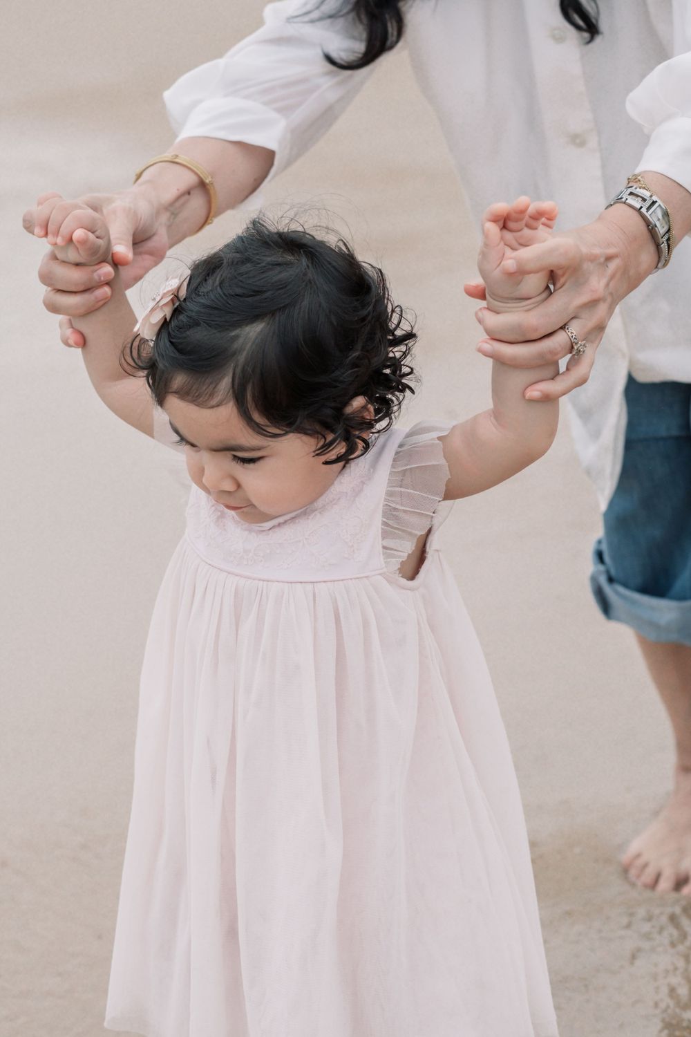 Small child in white dress holds hands with adult while standing on beach.