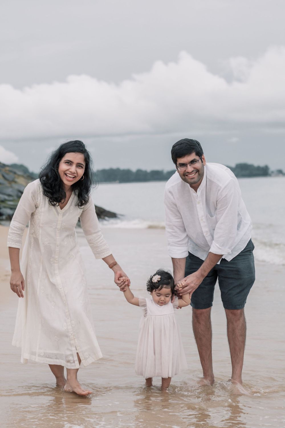 Family holds hands while standing in shallow ocean water wearing white outfits.