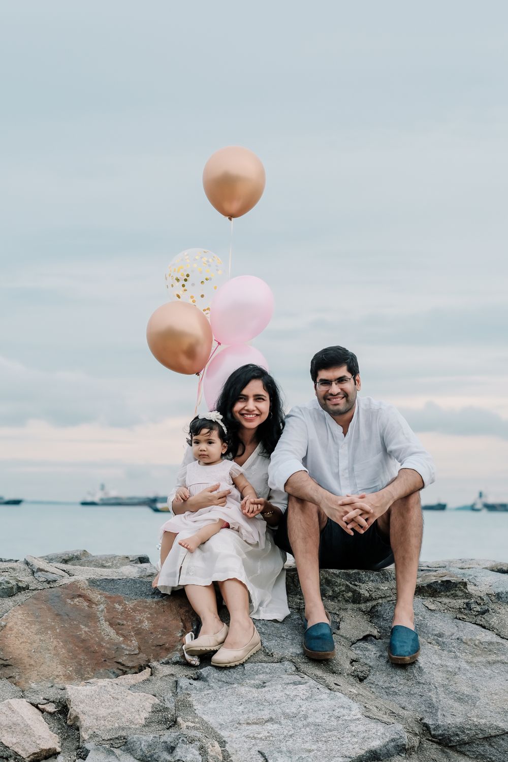 Family sits on rocky shore with pink and gold balloons against ocean backdrop.