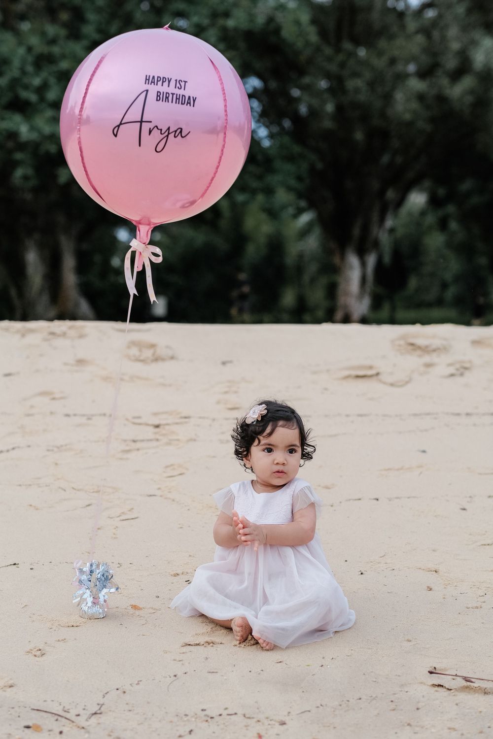 A toddler in a white dress sits on sand beneath a pink birthday balloon.