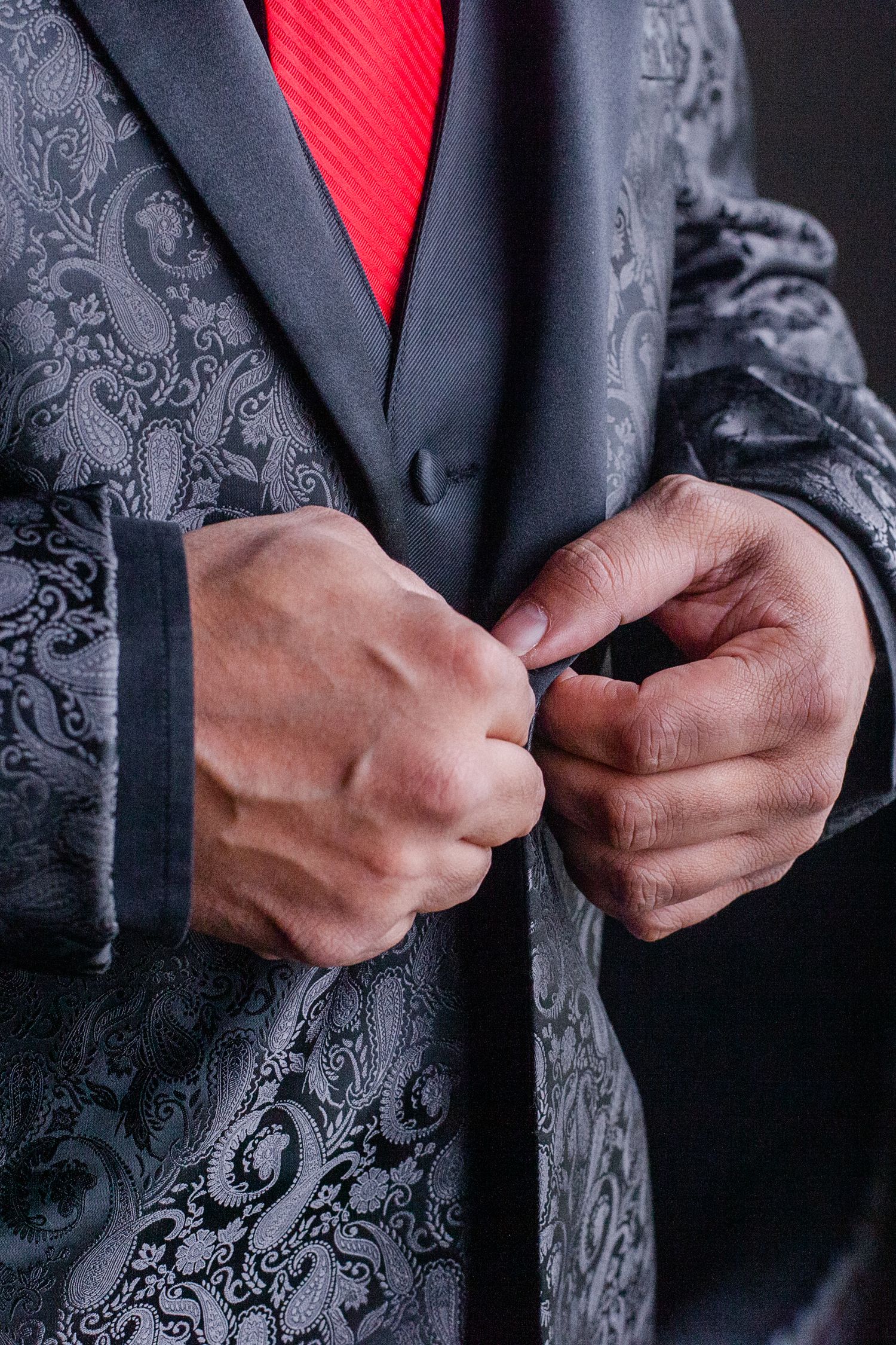 Close-up of hands buttoning ornate black and grey paisley suit jacket with red accent.